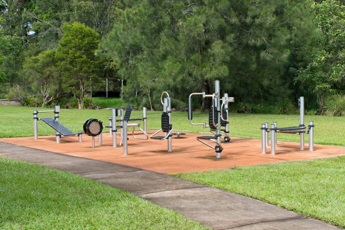 Outdoor exercise equipment on a concrete pad surrounded by grass and trees in a park setting.