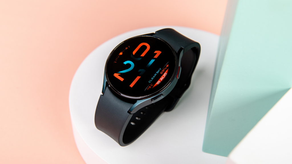 A black smartwatch with a colorful digital display, resting on a white circular platform against a soft pink and mint green background.