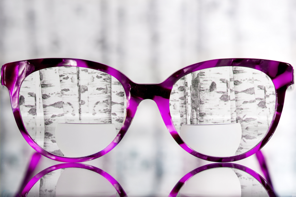 Purple cat-eye eyeglasses with patterned lenses placed on a reflective surface, showing an artistic image of trees through the lenses.