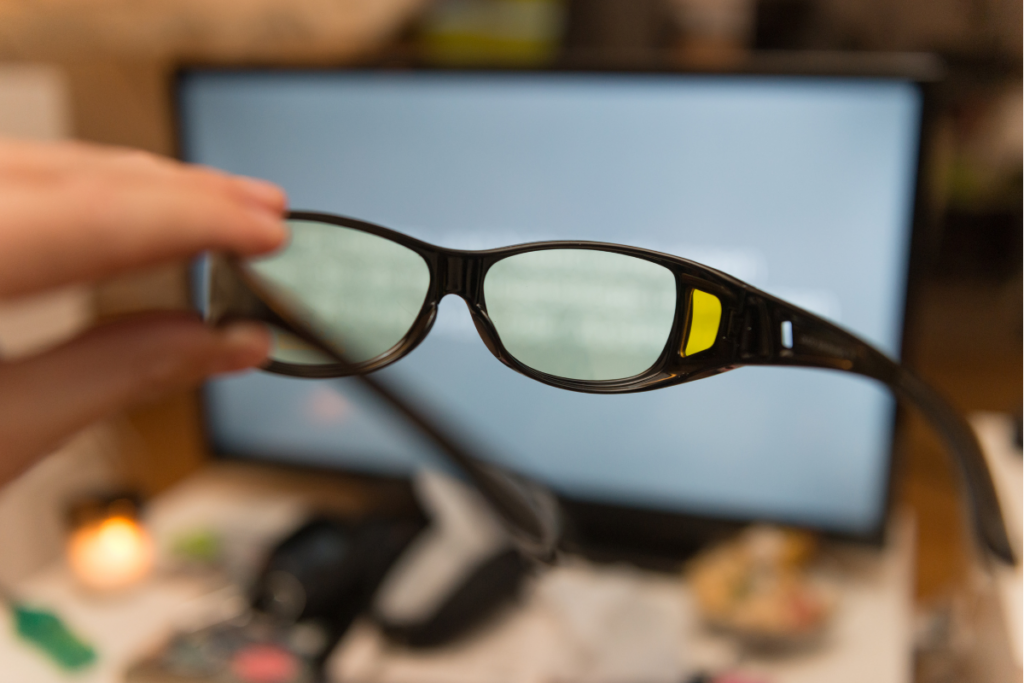 A hand holding a pair of dark-framed glasses with yellow-tinted lenses in front of a blurred computer screen.