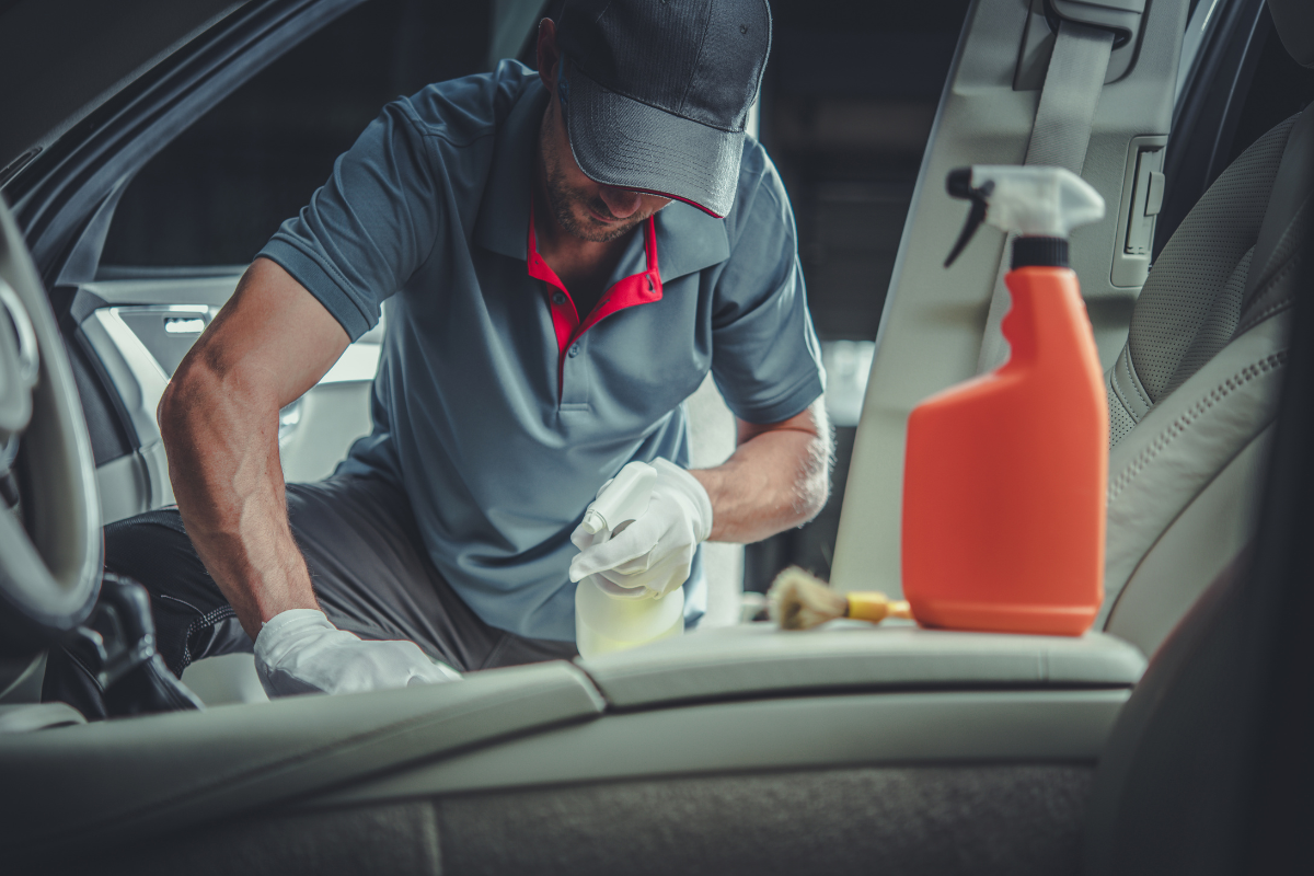 Man wearing gloves and a cap cleaning a car interior using a spray bottle, with an orange cleaning solution container placed on the seat.
