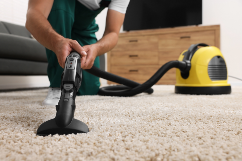 A person using a wet vacuum cleaner on a beige carpet, with a yellow vacuum unit in the background.