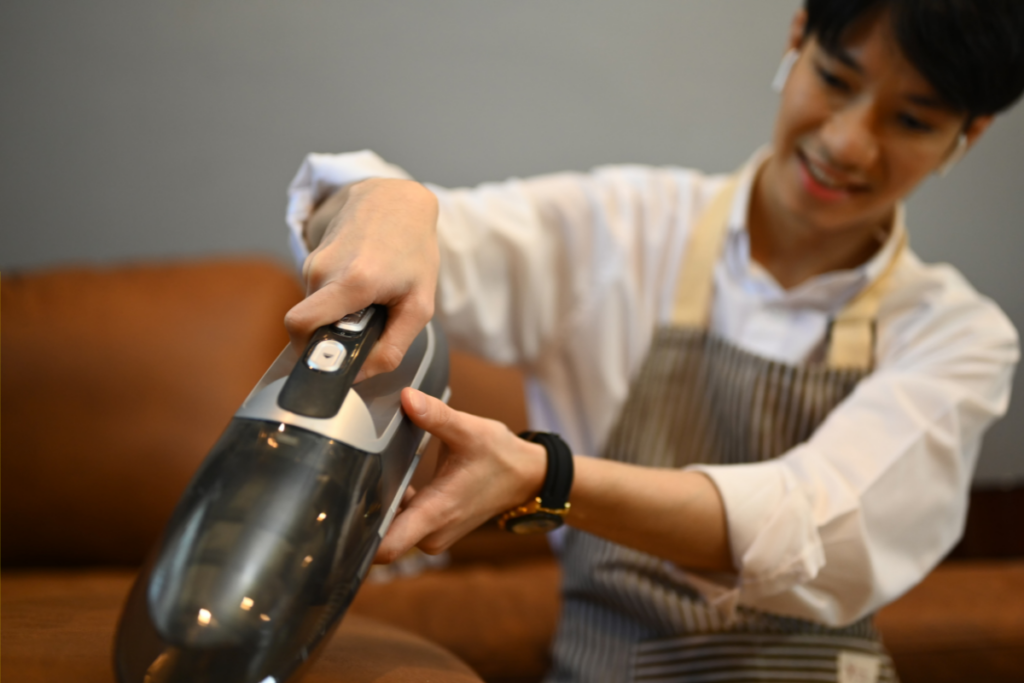 Person using a handheld vacuum cleaner to clean a couch while wearing a striped apron and smiling.