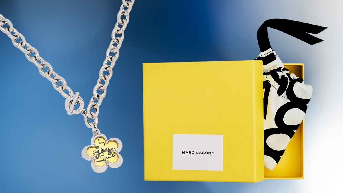 Marc Jacobs The Daisy Puzzle Necklace in silver with yellow daisy pendant, displayed beside the signature yellow Marc Jacobs gift box and patterned dust bag on a blue background.