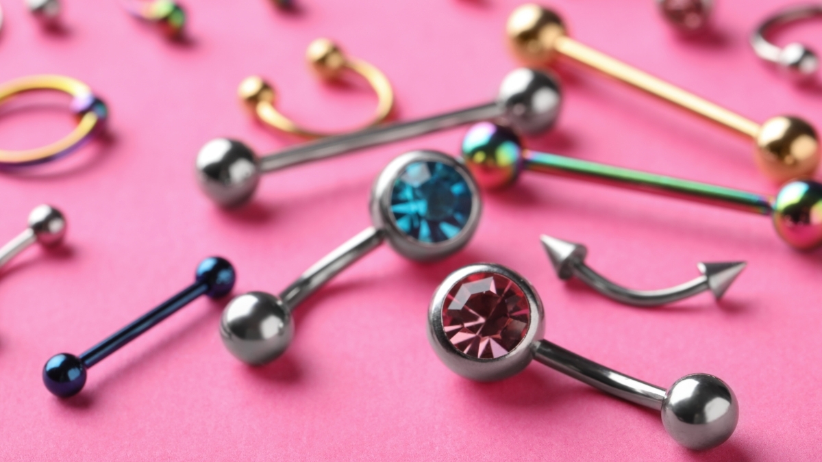 A close-up of colorful body piercing jewelry, including barbells, curved bars, and rings in gold, silver, and rainbow finishes, displayed on a pink background — perfect for illustrating Claire’s Piercing Guide and jewelry options.