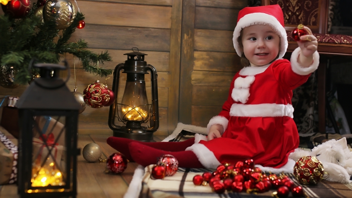 A smiling toddler dressed in a red Santa outfit sitting by a Christmas tree with ornaments, lanterns, and festive holiday decorations.
