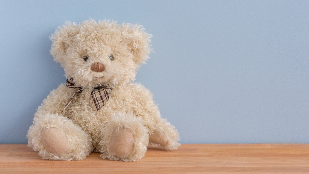 Soft beige teddy bear with a plaid bow sitting on a wooden surface against a light blue wall.