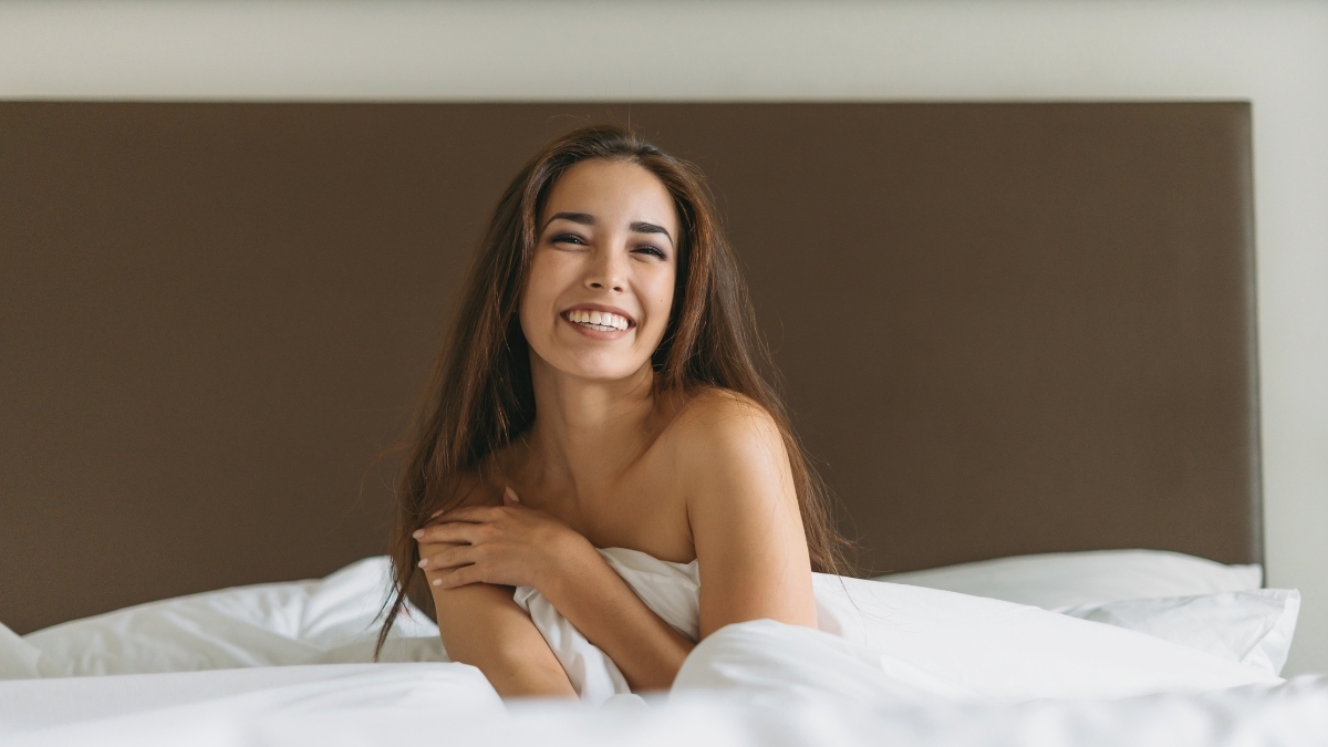 Woman sitting in bed smiling with natural, healthy skin and relaxed intimate wellness vibes.