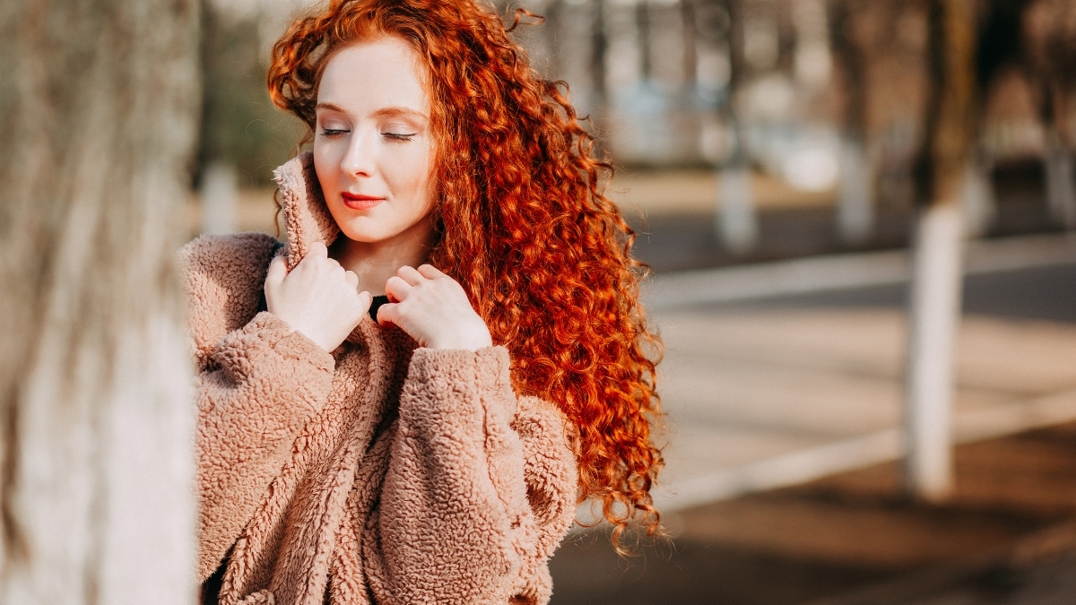 Woman with long curly red hair wearing a cozy tan fleece jacket outdoors during cold weather, showcasing warm and stylish winter layering.