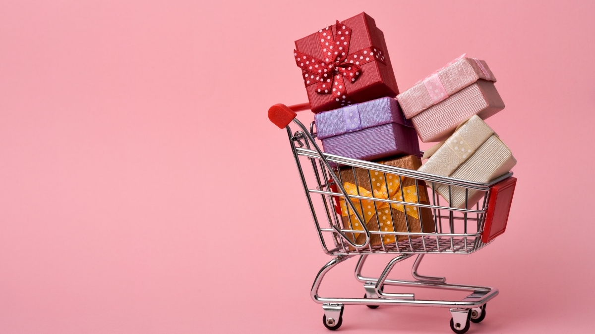 Colorful gift boxes stacked in a small shopping cart against a pink background, symbolizing holiday gift shopping and budget-friendly presents.