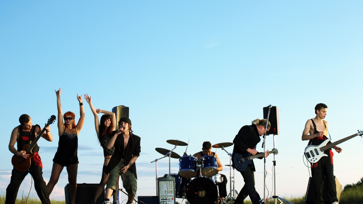 Outdoor rock band performing live on a sunny day with guitarists, vocalists, drummer, and audience members raising their hands.