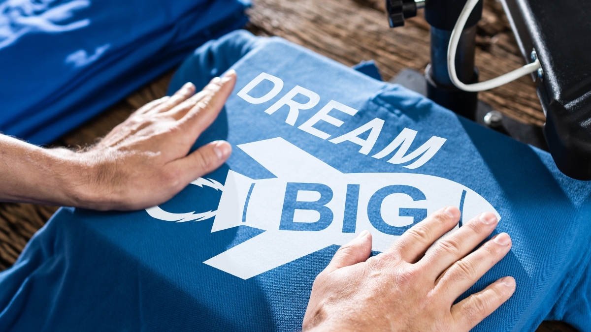 Hands pressing a custom heat-transfer design onto a blue T-shirt with the text “Dream Big” during the shirt printing process.