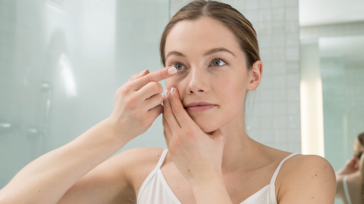 Woman inserting a contact lens in a bathroom mirror, demonstrating safe and proper contact lens application.