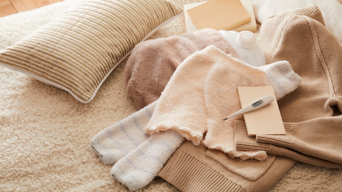 Cozy winter essentials including soft blankets, warm sweaters, fuzzy socks, and a digital thermometer arranged on a bed.