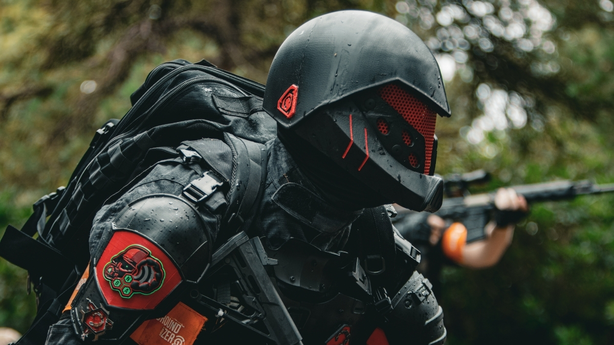 Tactical soldier in futuristic black armor and helmet with red accents carrying gear in an outdoor forest setting.