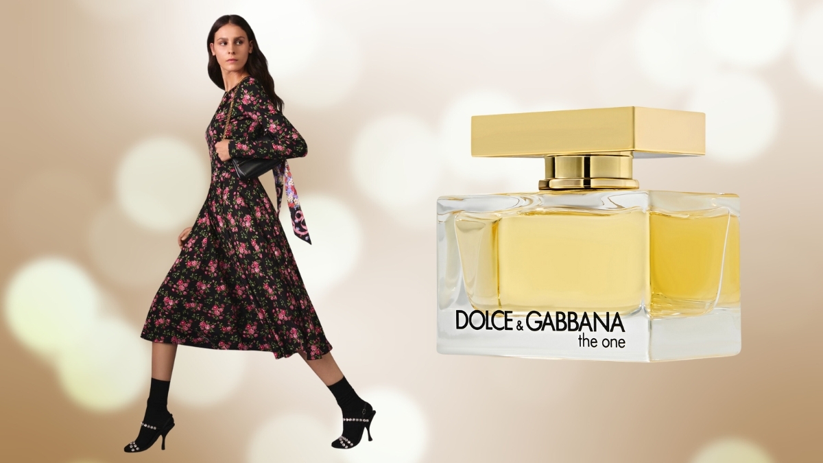 Woman wearing a black floral Dolce & Gabbana–style dress walking beside a large bottle of Dolce & Gabbana The One perfume against a soft, golden bokeh background.