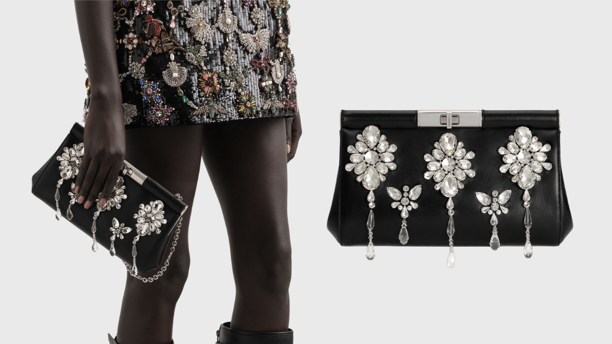 Black evening clutch with crystal floral embellishments and dangling jeweled accents, shown held by a model wearing a beaded mini dress.